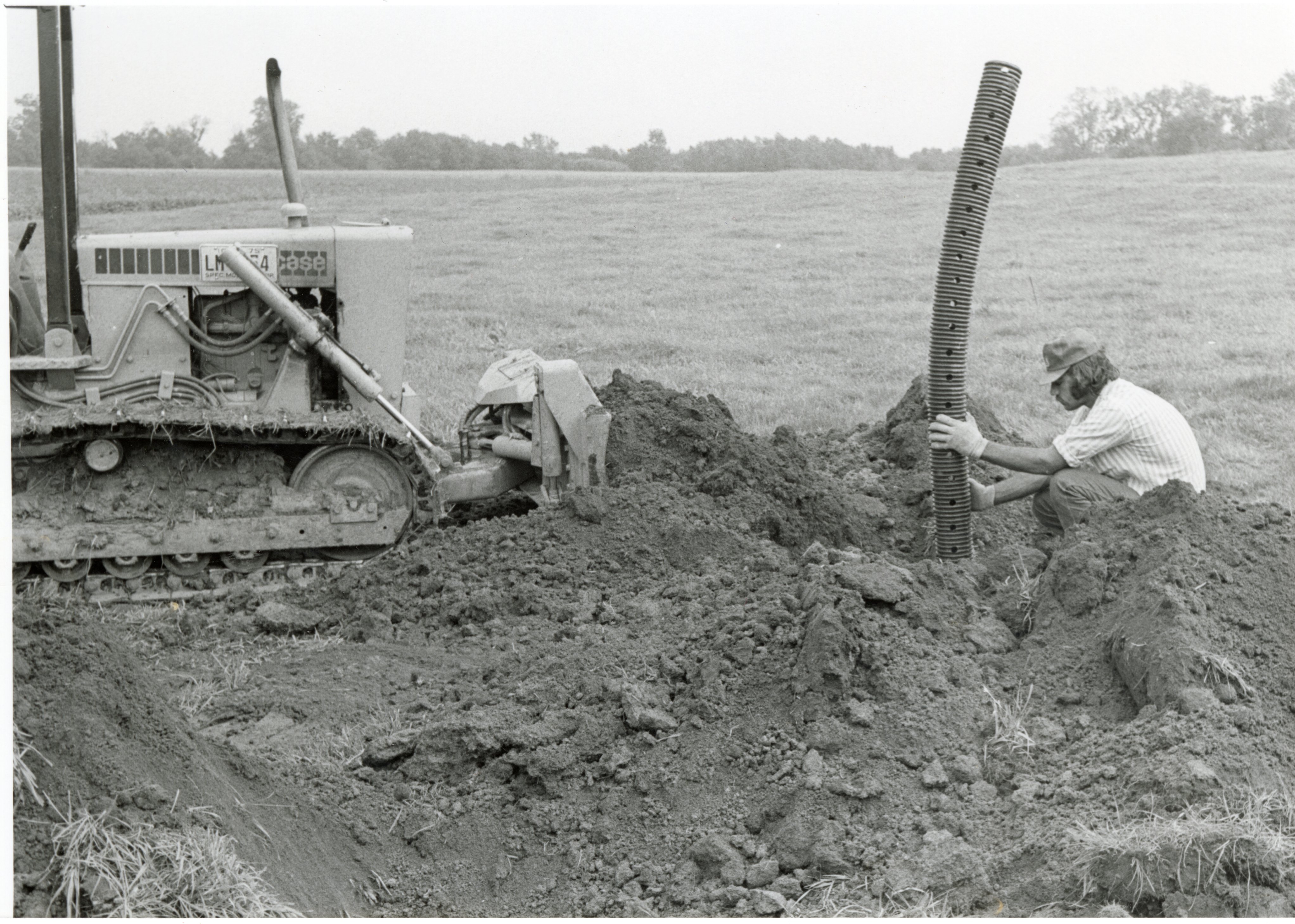 1979  Man on Bulldozer in front of Man putting pipe in the ground