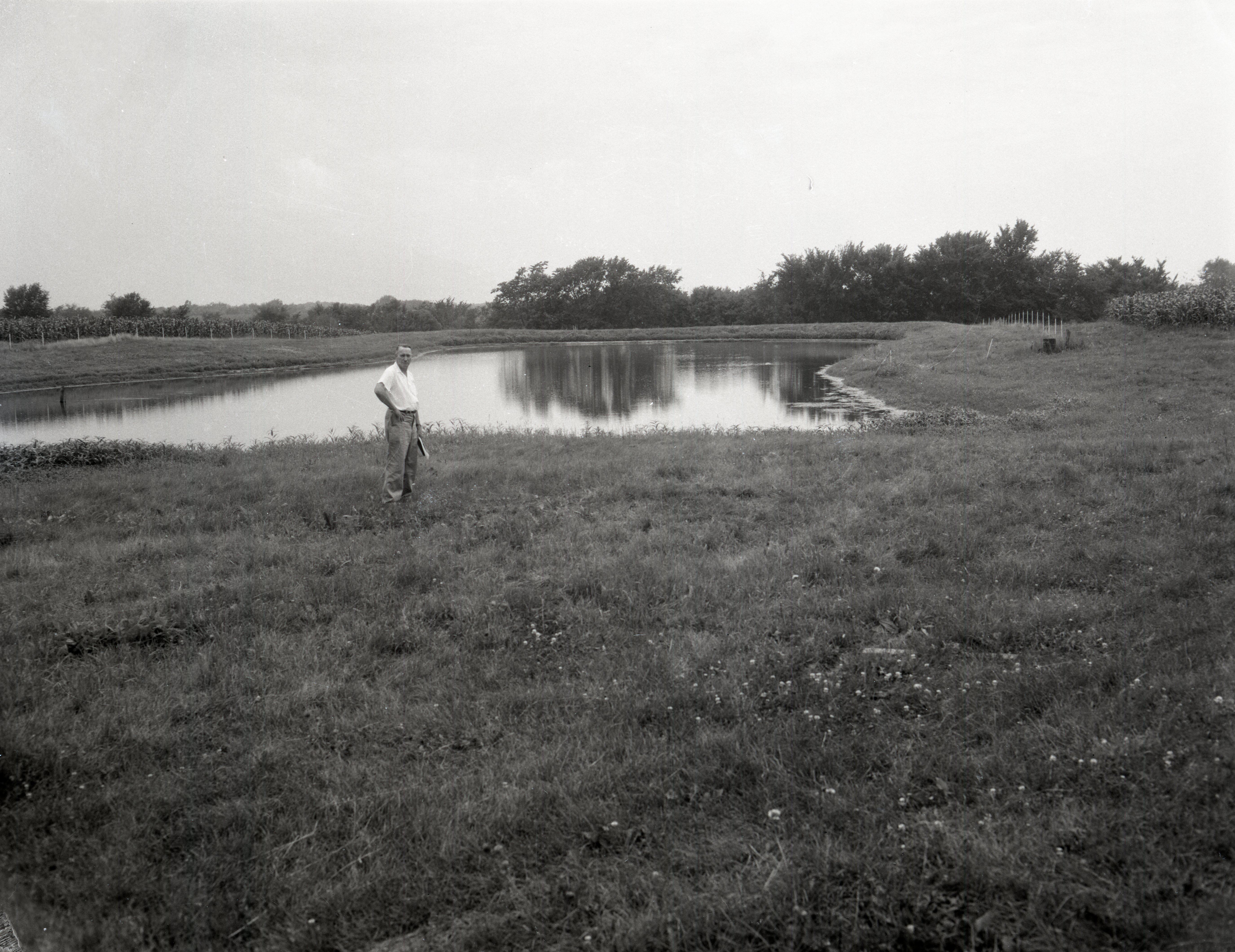 Man stands by farm pond holding hat