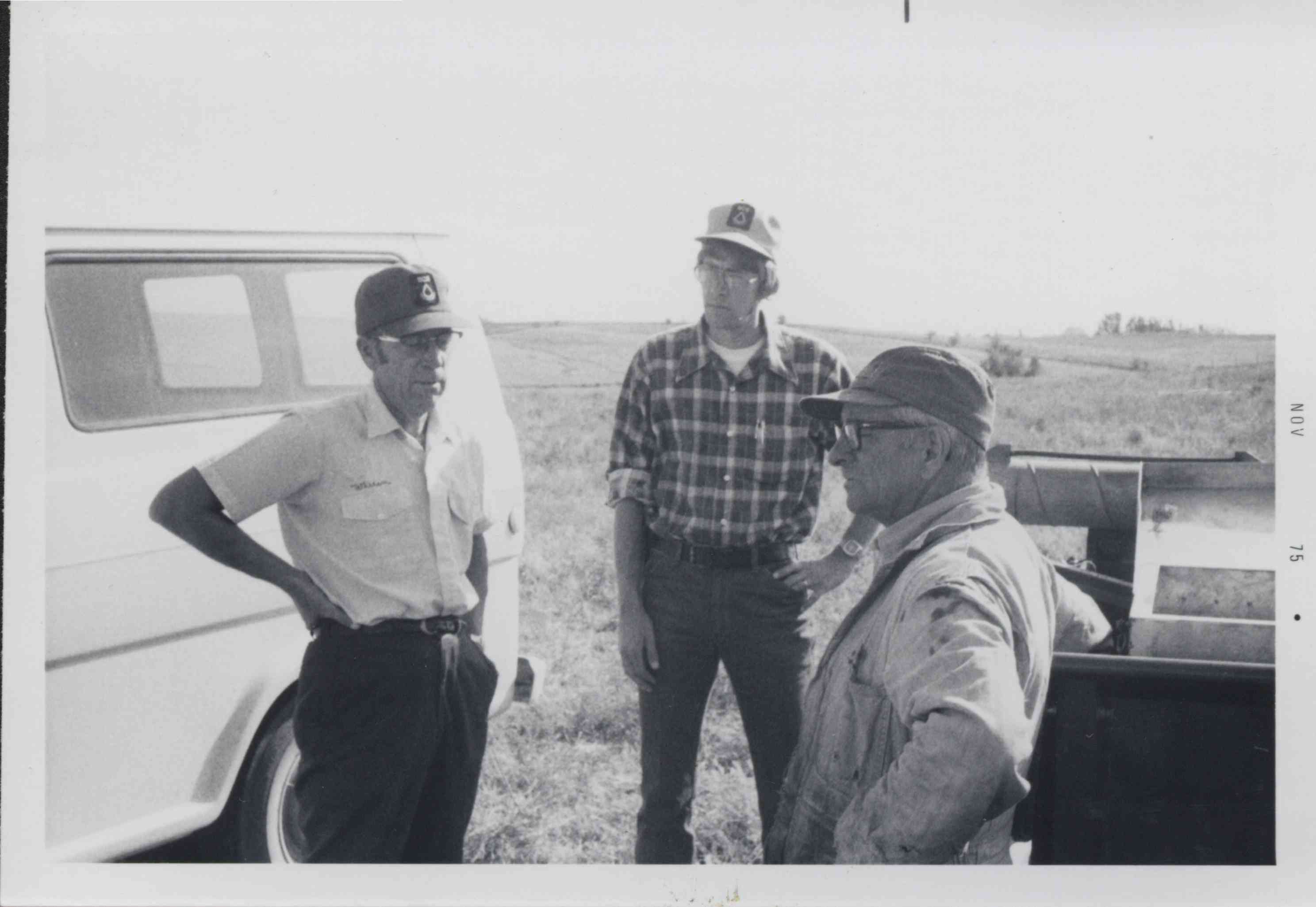 Three Men Standing in Field by Van