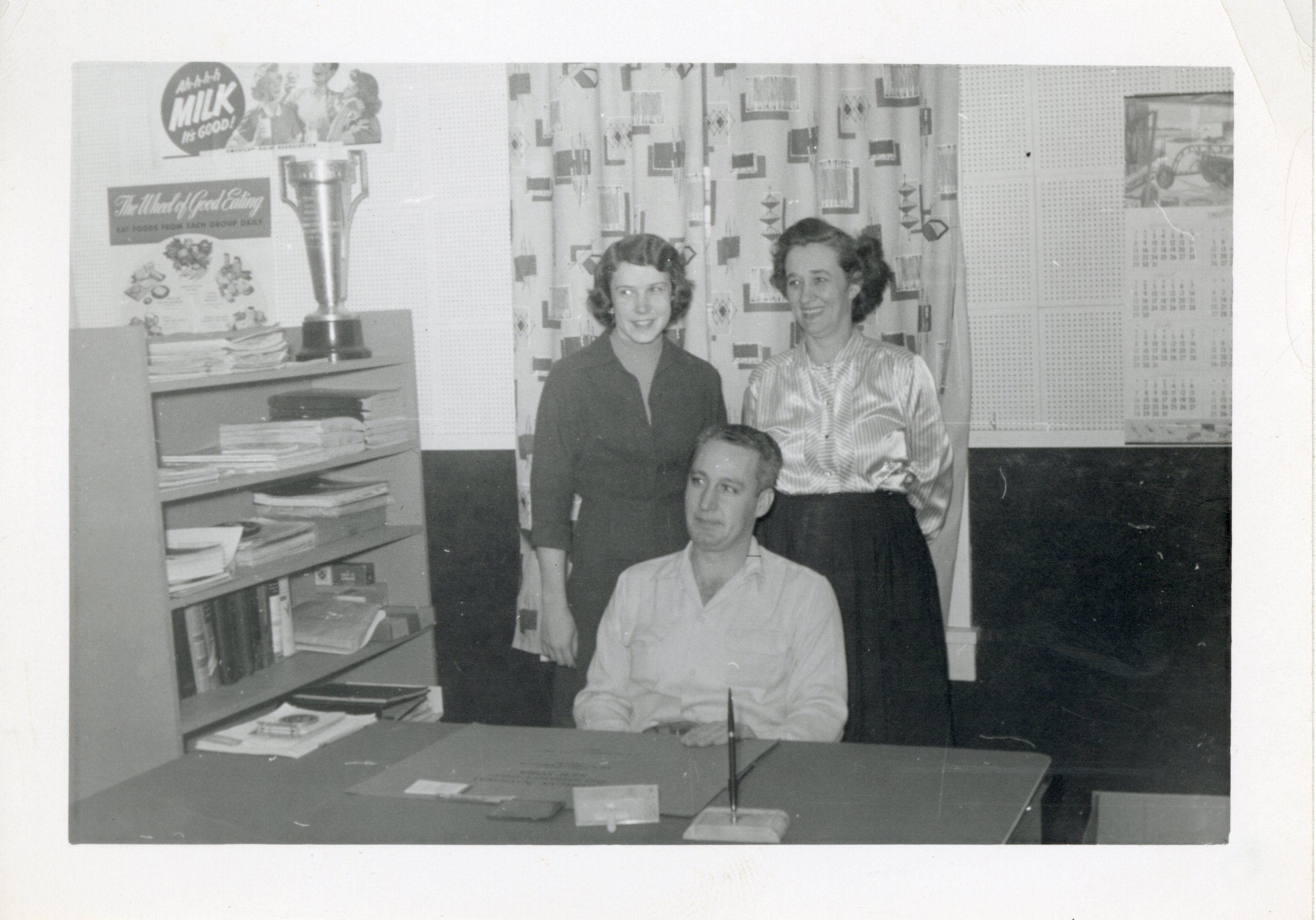 Bill Byers at desk with two women standing behind