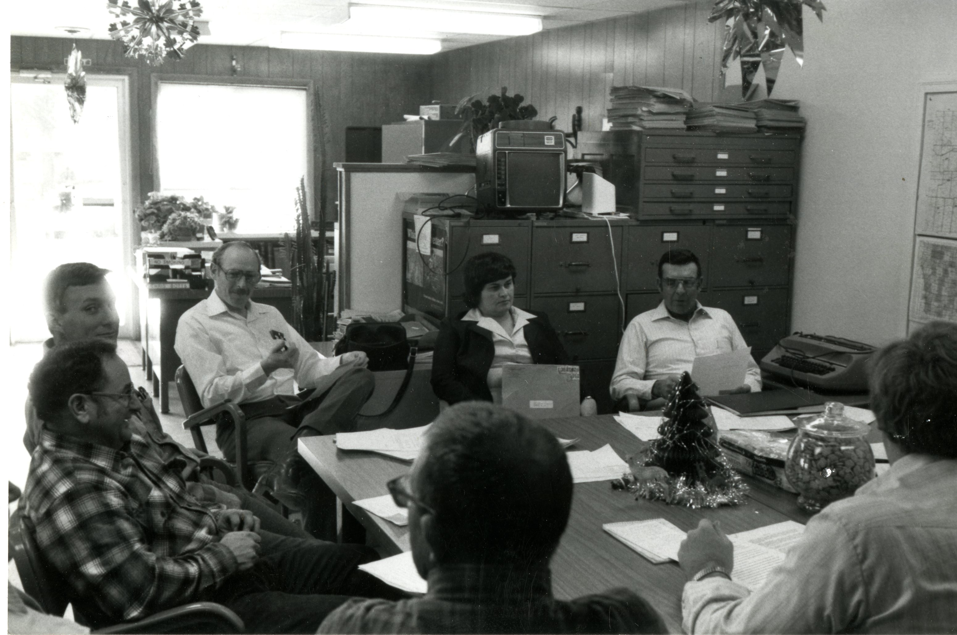 Six men and one woman meet around a table for a soil conservation meeting in December 1984
