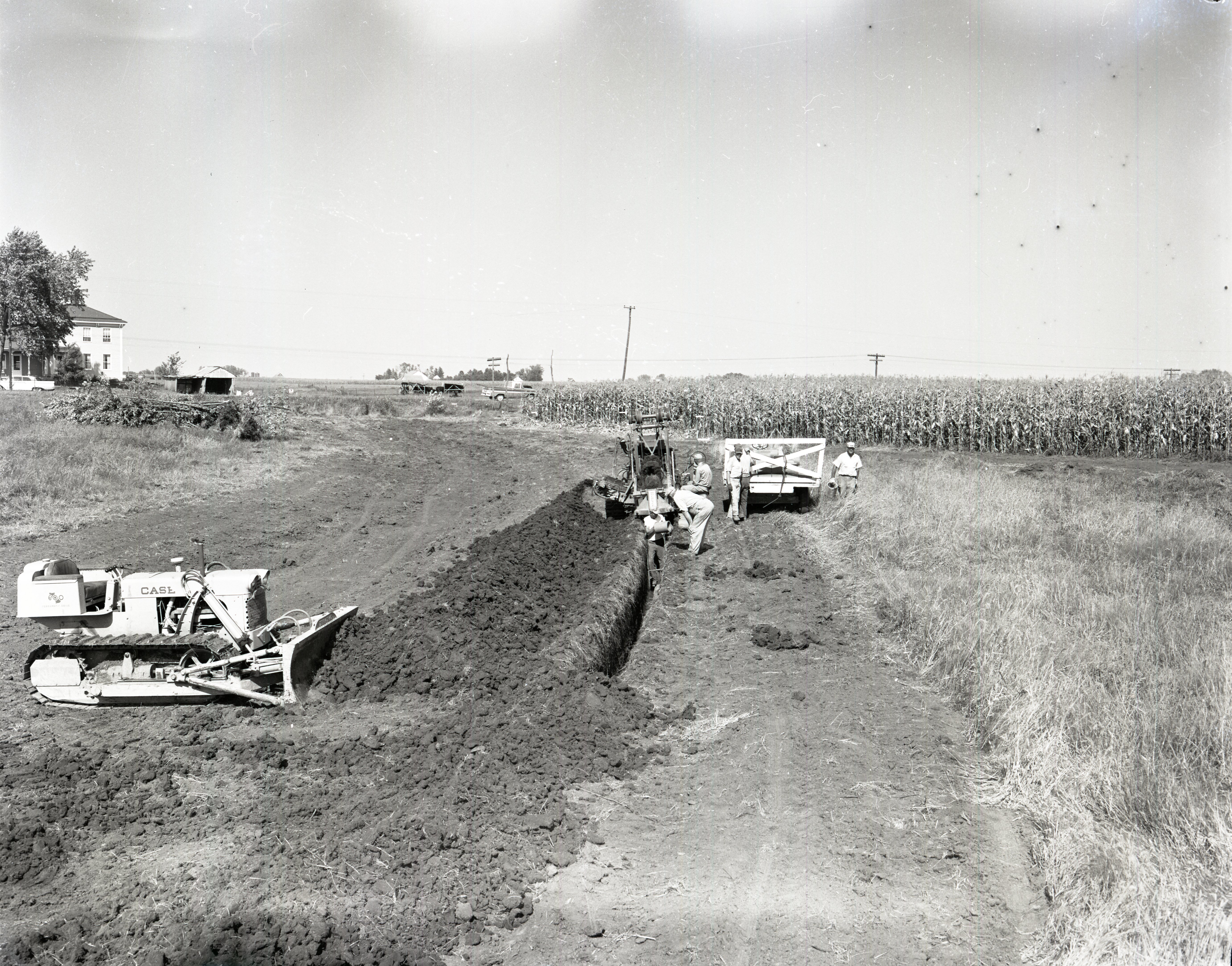 Man holding pipe stands in trench in front of trench digger near cornfield