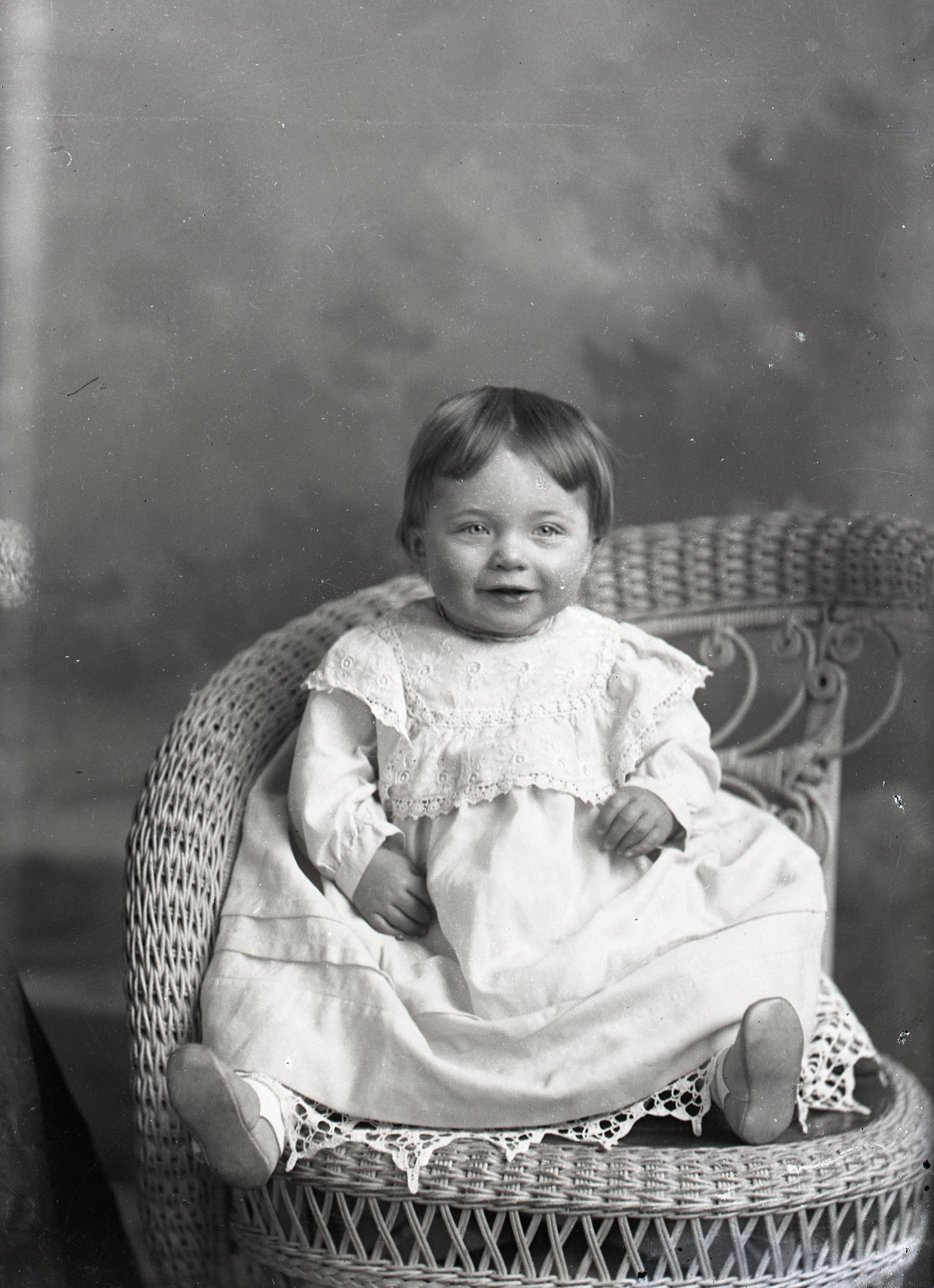 Infant smiling in white dress, seated in wicker chair