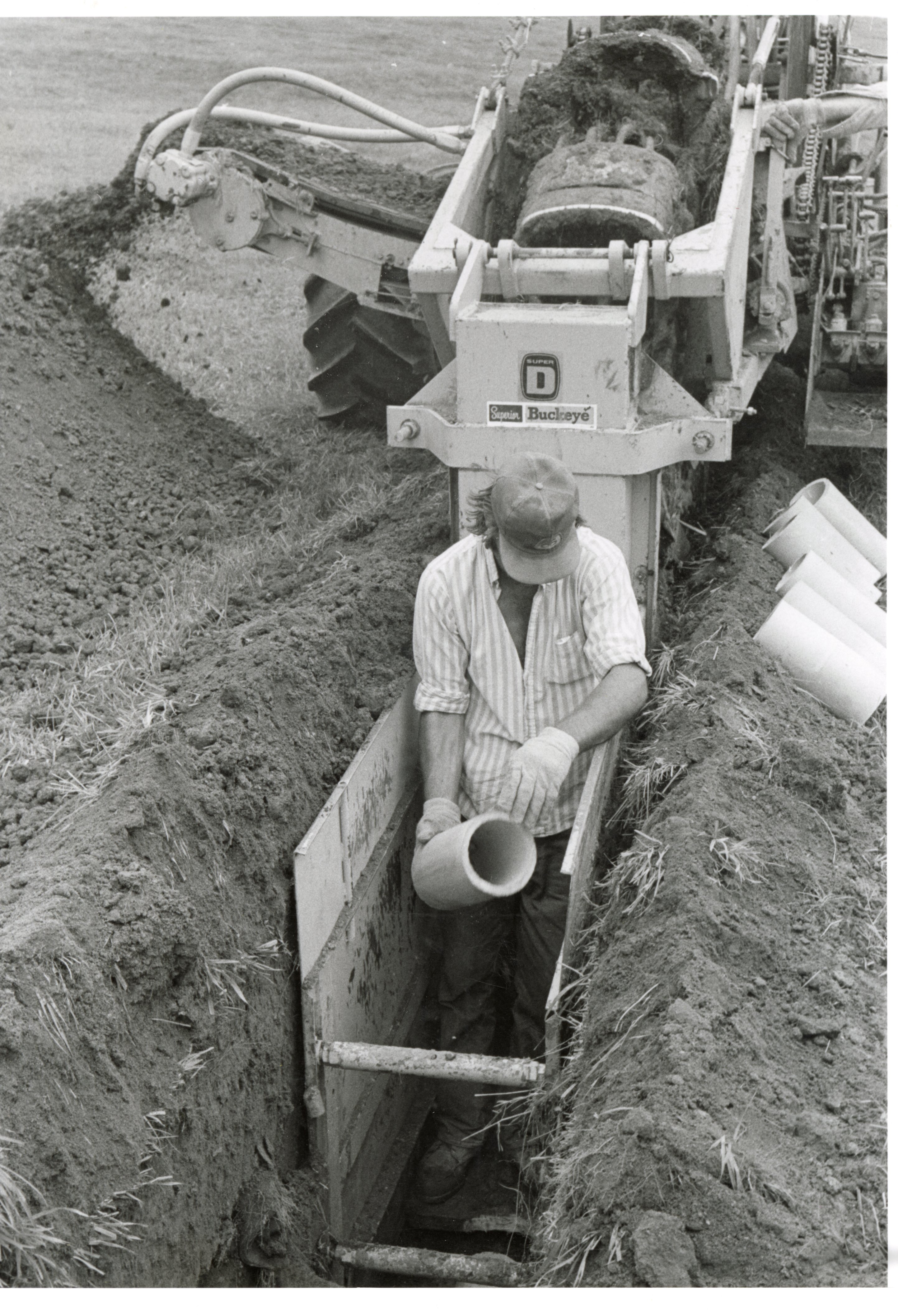 1979  Man holding pipe stands in trench in front of trench digger