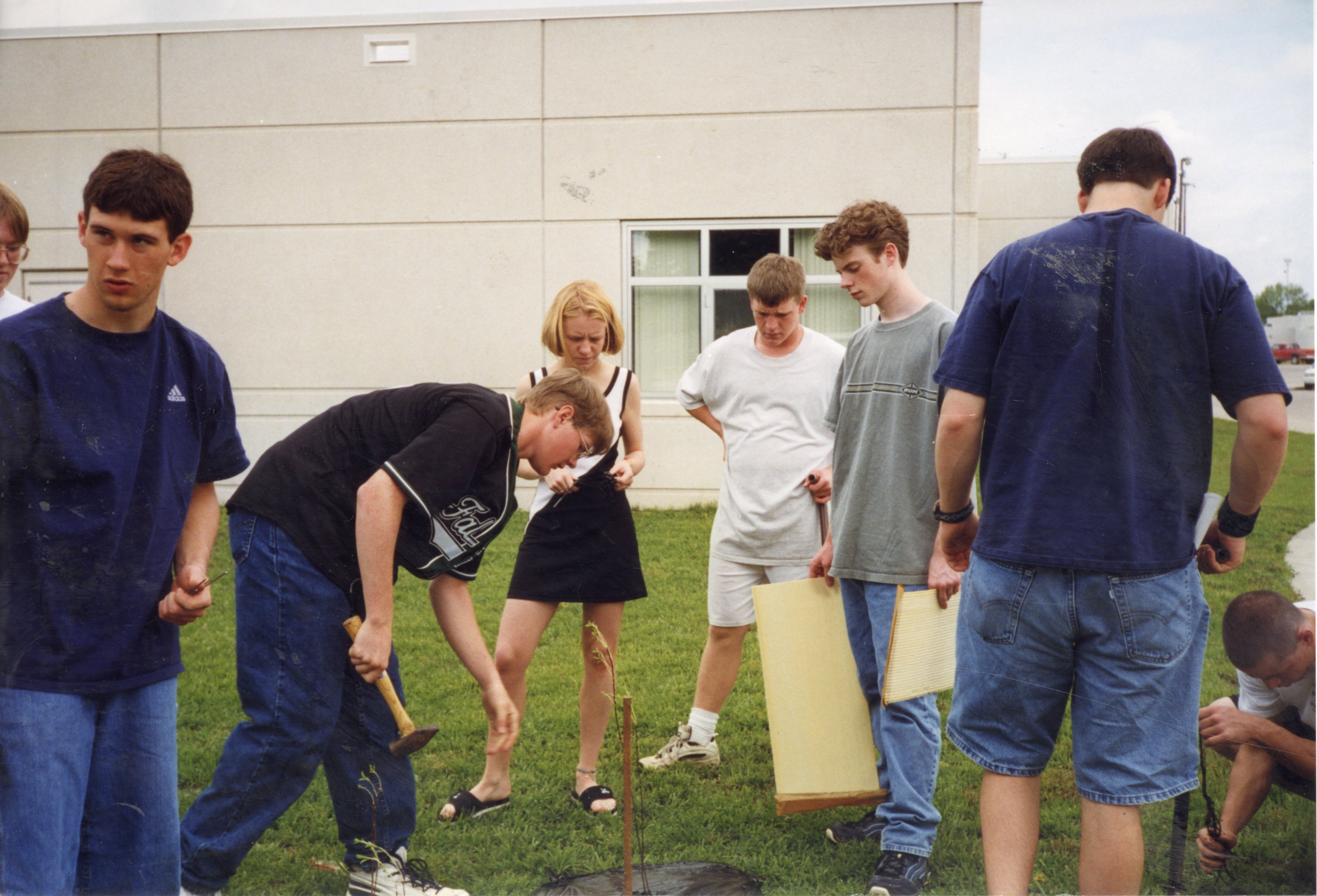 1999 Students watch Ben Schwenk drive a stake into the ground in preparation for planting a tree while students watch