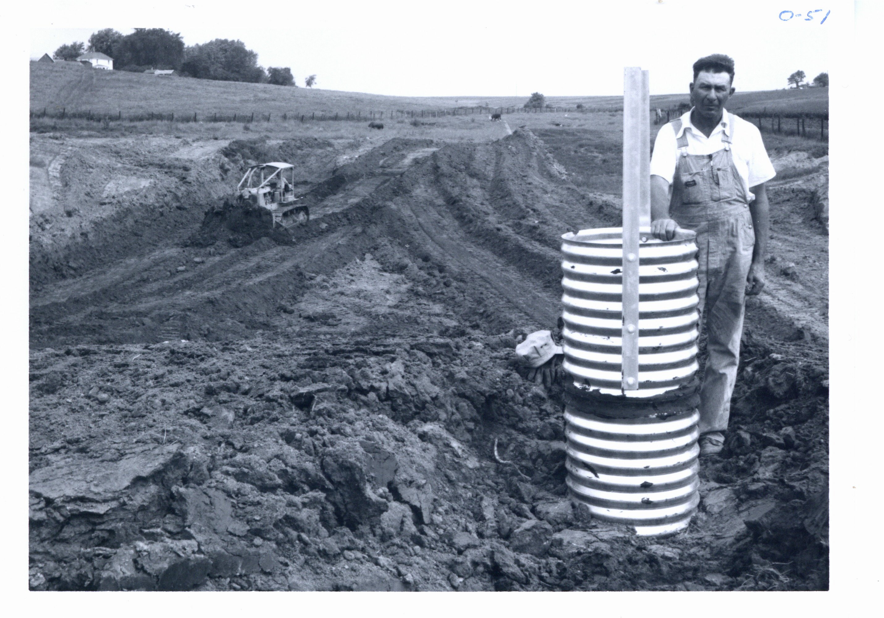 Clyde Small stands beside inlet of pipe spillway of his pond under construction, 1966