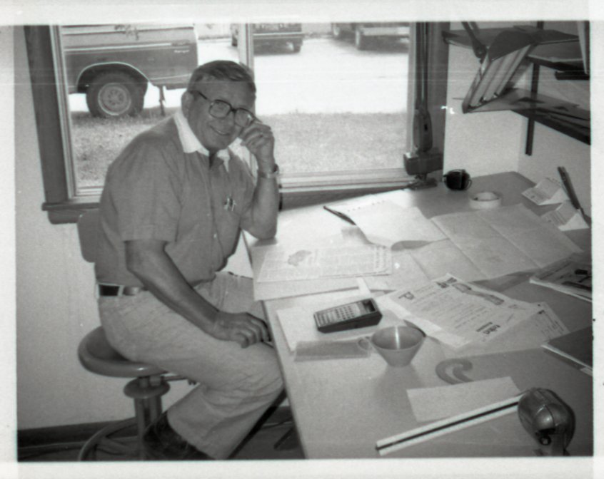 Unidentified Smiling Man with Glasses Sits at Desk in Office