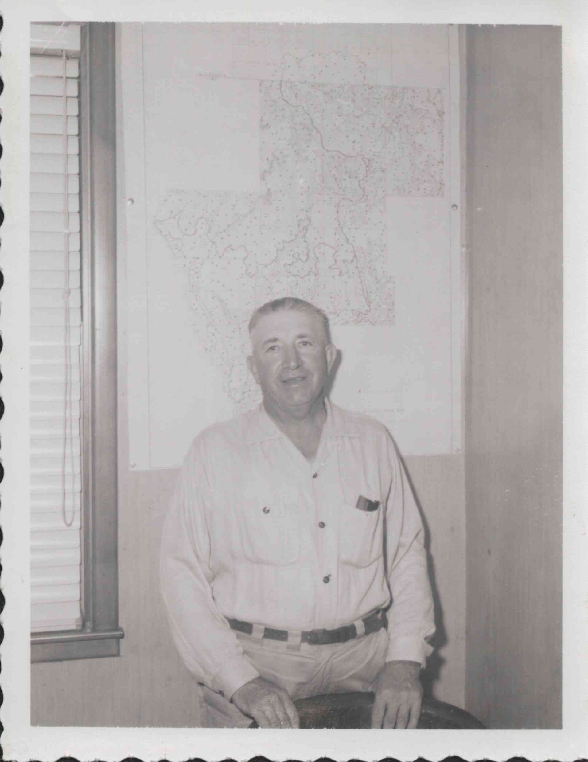 Unidentified Man Holding onto Back of Chair with Map on Wall behind him