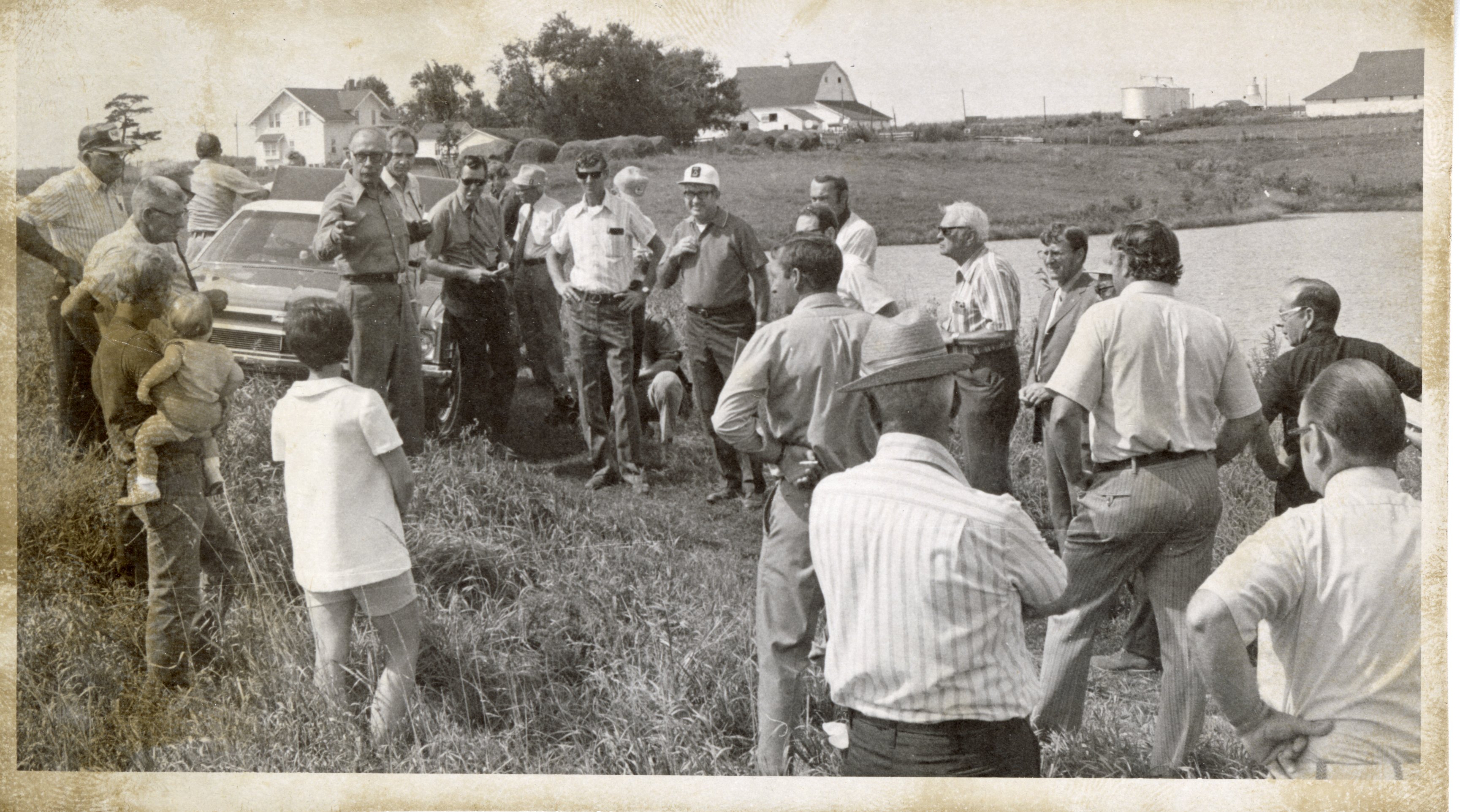 Unidentified Man with Glasses Talks to Group Standing Beside Farm Pond