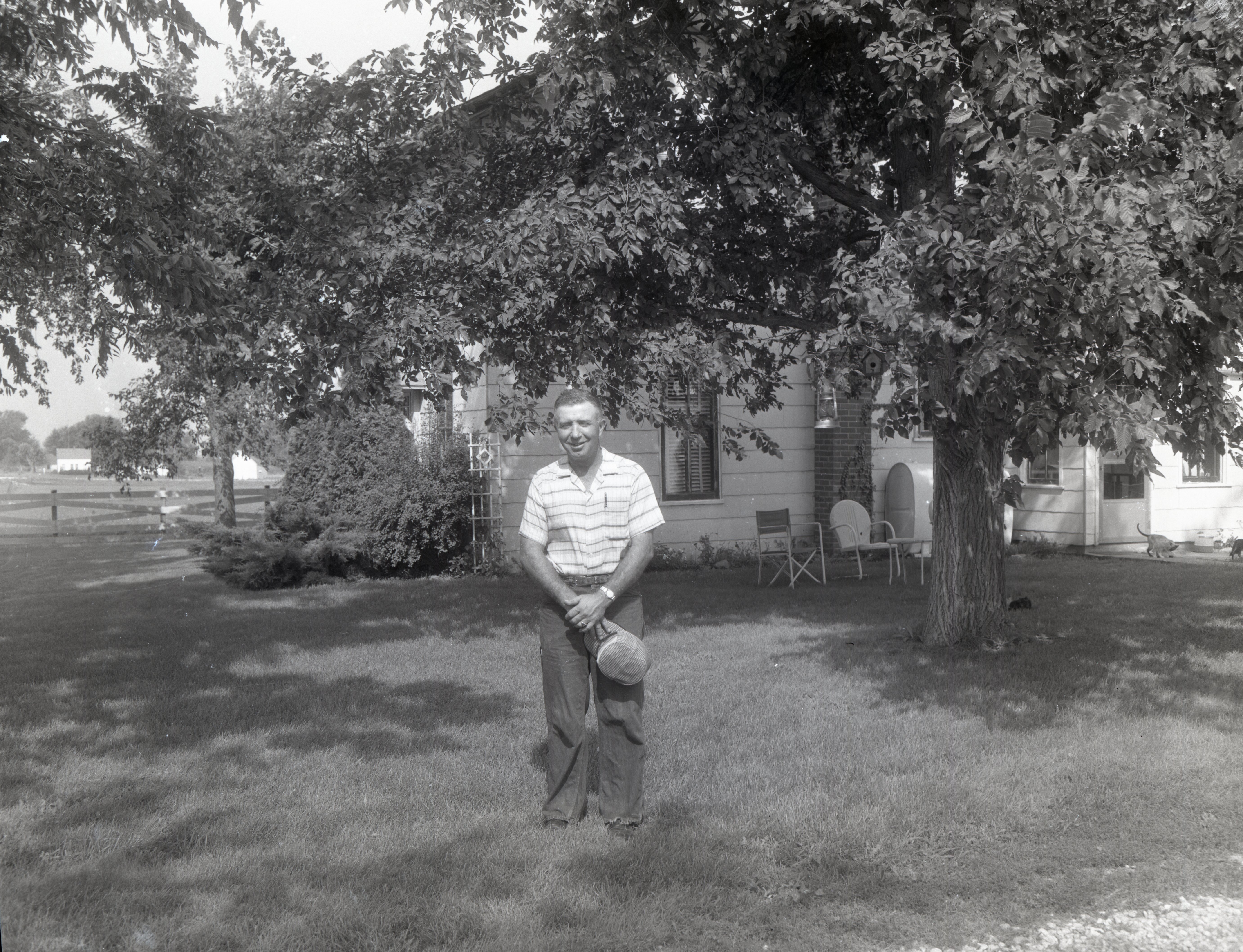 Unidentified man holding hat stands in front of farmhouse