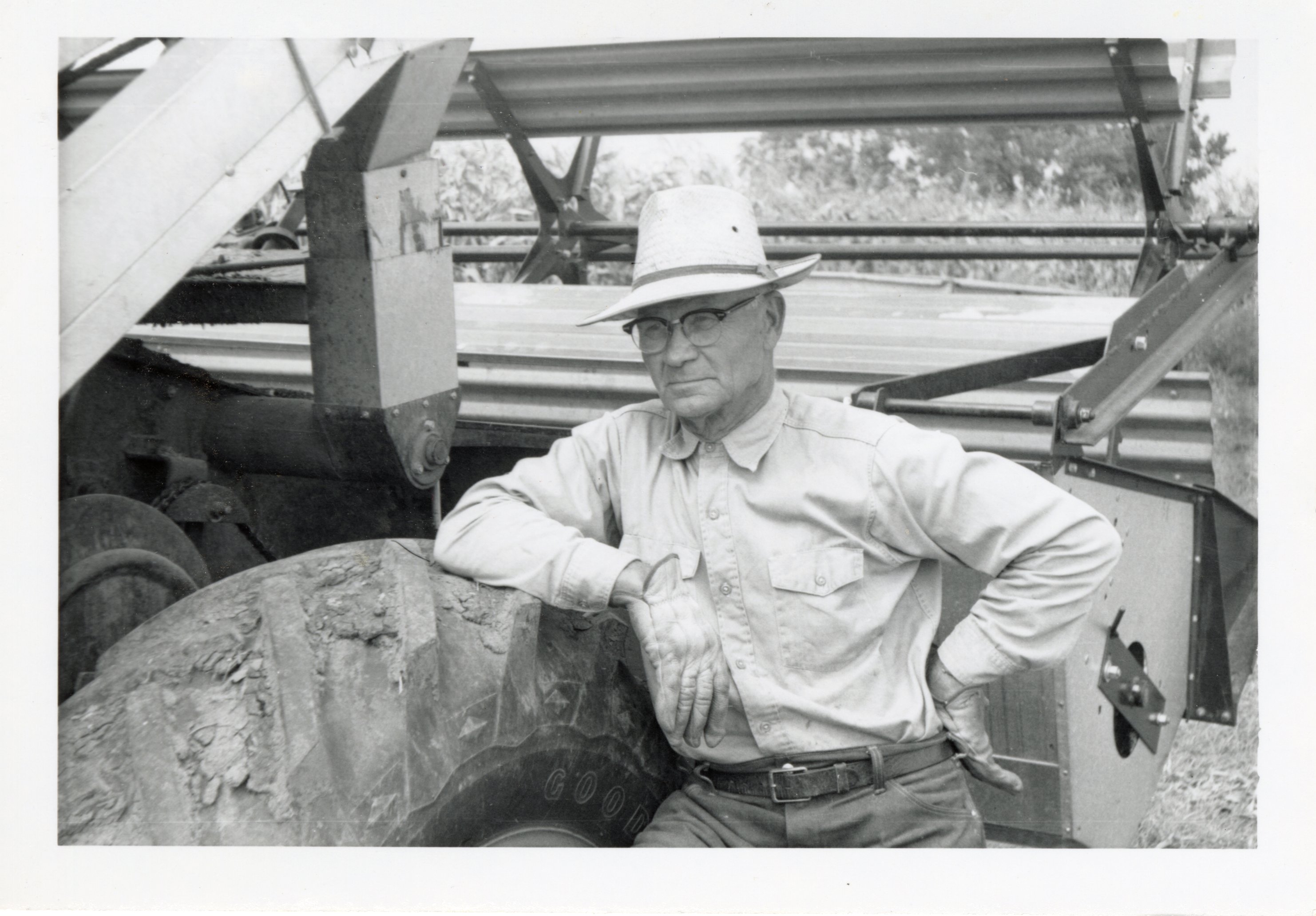 1964 _ Man with glasses and hat stands beside farm machinery