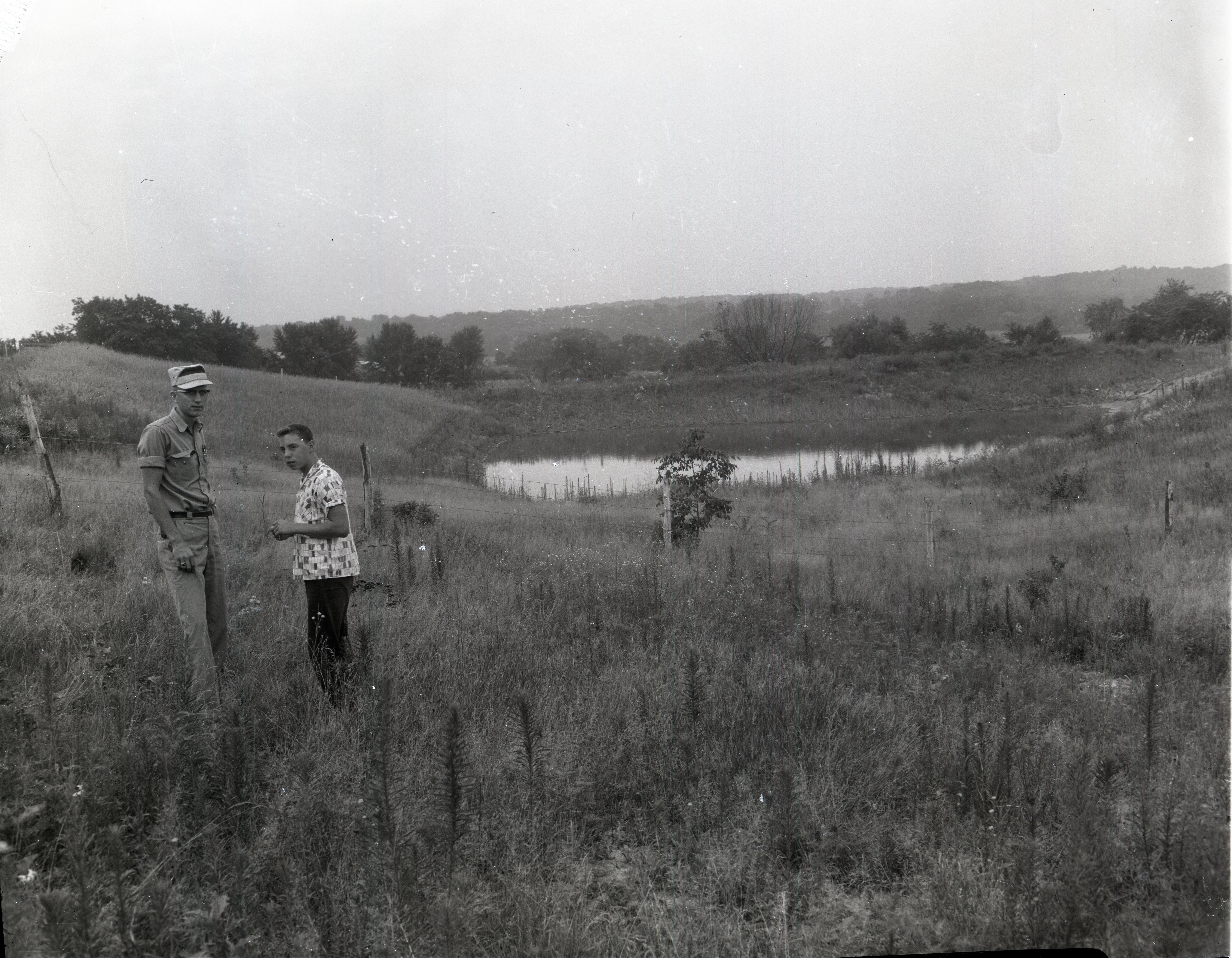 Two unidentified men stand in field by pond