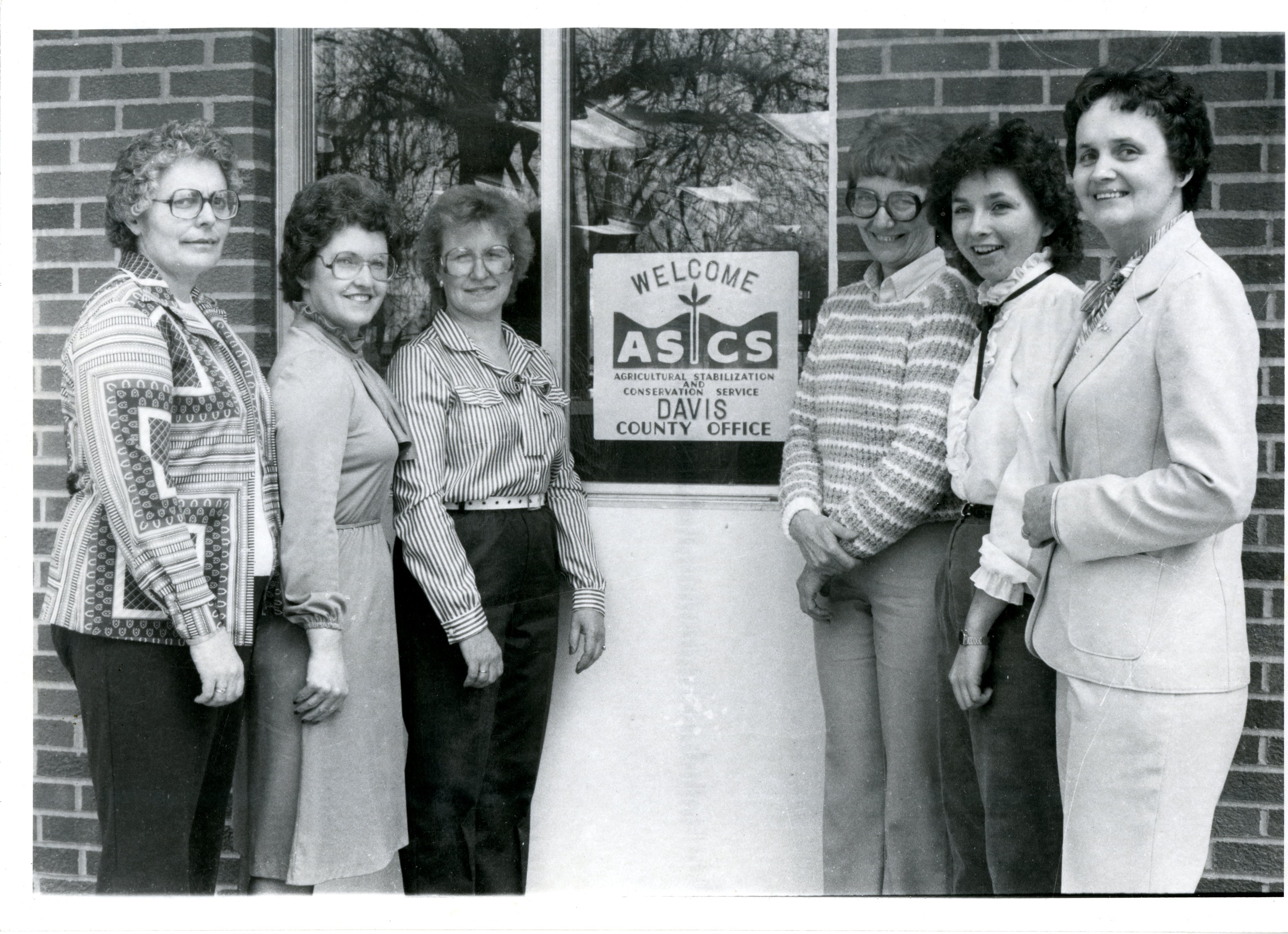 Six women stand on either side of a welcome sign for the Agricultural Stabilization Conservation Service,<br />
 Davis County Office