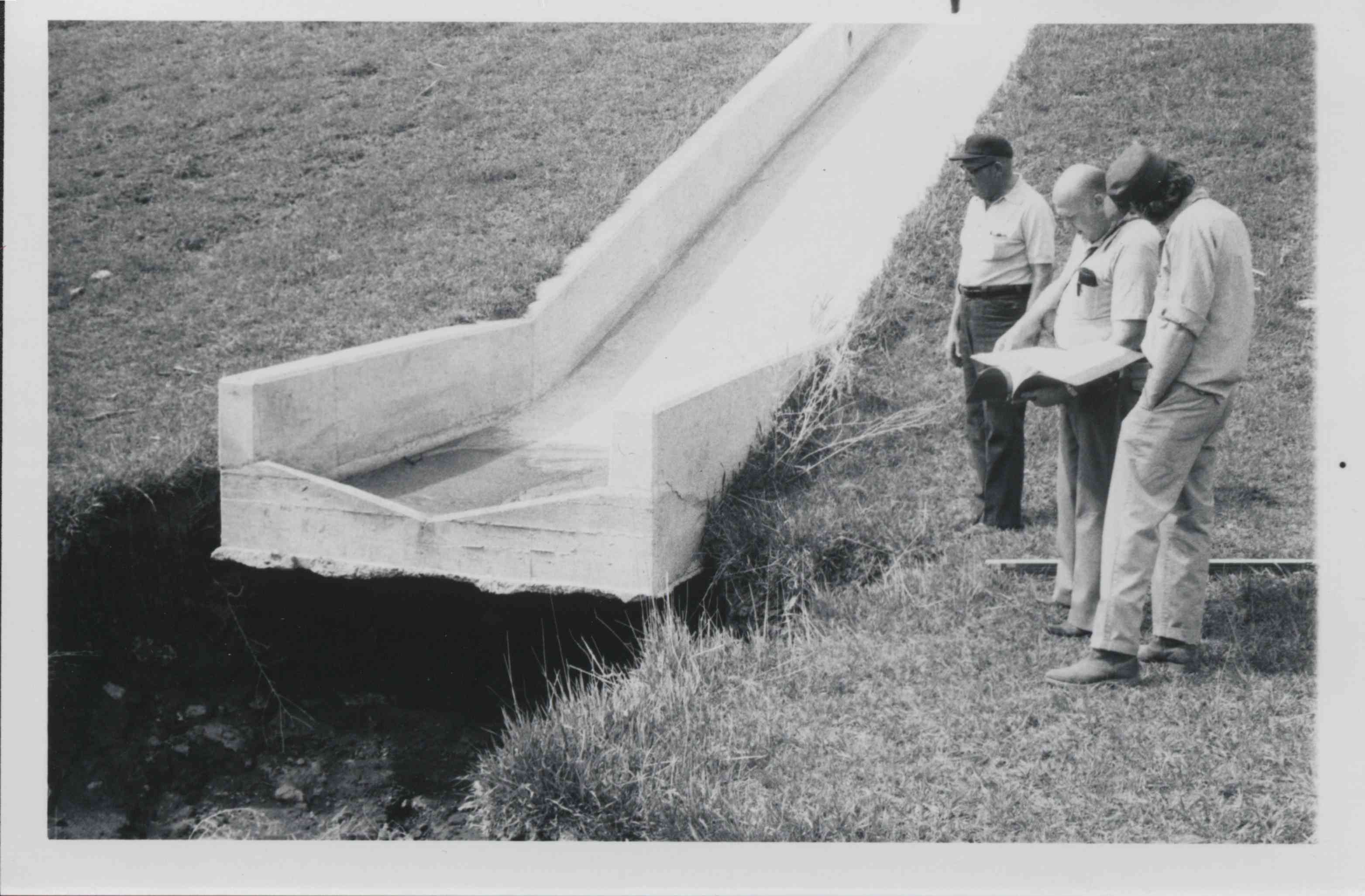 Three men stand by water drainage culvert looking at book