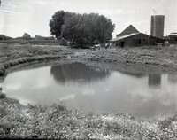 Two unidentified men stand with horses beside a barn in back of a pond