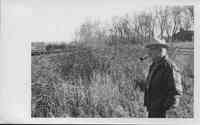 Man with white goatee in white hat wearing glasses and smoking a pipe stands in field by tall grass.