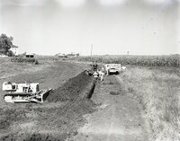 Man holding pipe stands in trench in front of trench digger near cornfield