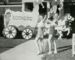 Two girls are posed in front of a covered wagon lawn display at Homecoming, 1946
