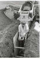 1979  Man holding pipe stands in trench in front of trench digger