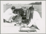 Two researchers taking measurements with equipment in a field, with pigs in the background.