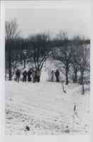 Group of unidentified men and couple standing in the snow at the top of an enbankment by trees looking at some papers