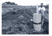 Clyde Small stands beside inlet of pipe spillway of his pond under construction, 1966