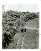 Farmer in overalls and glasses with hands in pockets stands next to shrubs