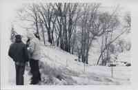 Three men stand in the snow looking at some papers