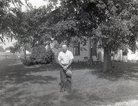 Unidentified man holding hat stands in front of farmhouse