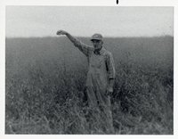 Farmer Wearing Overalls, Hat, and Glasses Stands in  Grassy Field with Arm Extended Above Head